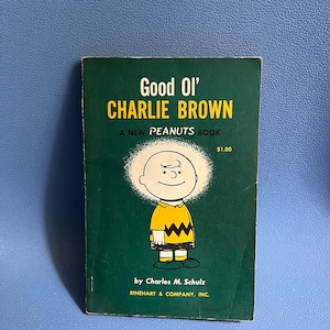Könnte beinhalten: Ein grüner Buchumschlag mit dem Titel "Good Ol' Charlie Brown" und einem neuen Peanuts-Buch. Der Umschlag zeigt eine Comic-Illustration von Charlie Brown mit einem gelben Shirt und einem schwarz-weißen Zickzackmuster.