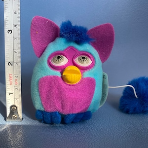 以下が含まれることがあります： 大きな目と黄色いくちばしを持つ青と紫のふわふわのおもちゃ。おもちゃは、ふわふわのポンポンが付いた青い尻尾を持っています。おもちゃは約15cmの高さです。