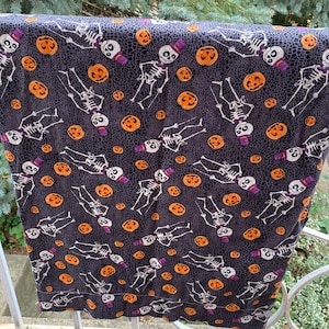 May include: A fabric textile featuring a repeating pattern of white skeletons wearing purple top hats and bow ties, interspersed with orange jack-o'-lanterns on a dark charcoal gray background with a fine, web-like black crackle texture.