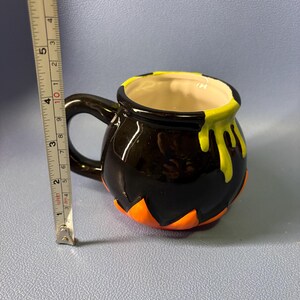 May include: A black ceramic Halloween cauldron mug with a handle. The rim has yellow drips, and the base has orange accents. The mug is approximately 4 inches tall.
