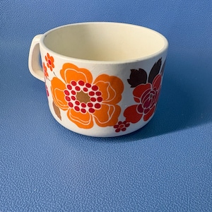 May include: A white ceramic mug featuring a vibrant floral pattern with large orange, red, and brown flowers. The mug has a rounded body and a single side handle, showcasing a bold, colorful design that wraps around the exterior of the vessel.
