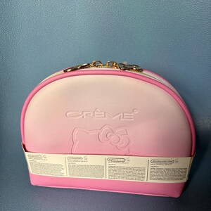 May include: A pink and white Hello Kitty-themed makeup bag with the word "CREME" embossed on the front. The bag has a zipper closure and a gold chain attached to the zipper pull. The bag is sitting on a blue surface.