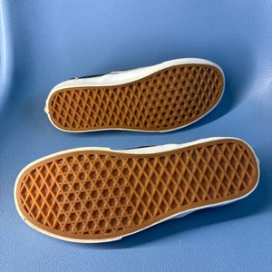 May include: Two black and white sneakers with brown rubber soles are displayed against a blue background. The soles feature a diamond pattern for traction. The shoes are positioned with the soles facing up, showcasing the design.