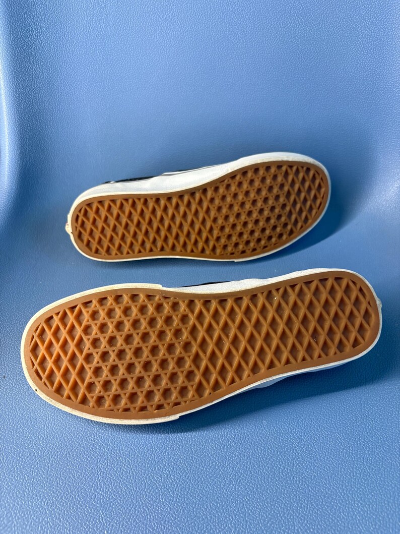 May include: Two black and white sneakers with brown rubber soles are displayed against a blue background. The soles feature a diamond pattern for traction. The shoes are positioned with the soles facing up, showcasing the design.