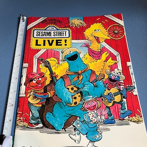 以下が含まれることがあります： セサミストリートライブ！のヴィンテージポスター。クッキーモンスター、ビッグバード、その他のキャラクターが赤い納屋の前で楽器を演奏しています。ポスターには「SESAME STREET LIVE！」と書かれており、Joe Mathiewのサイン入りです。