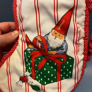May include: Christmas stocking with a white background and red vertical stripes. The stocking features a gnome illustration with a red hat and a white beard, wrapping a green polka-dotted gift box with a red bow. Candy illustrations are also present.