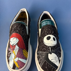 May include: Pair of black slip-on sneakers featuring characters from The Nightmare Before Christmas. One shoe displays Sally, the other Jack Skellington. Both shoes have white soles and a black patterned background.