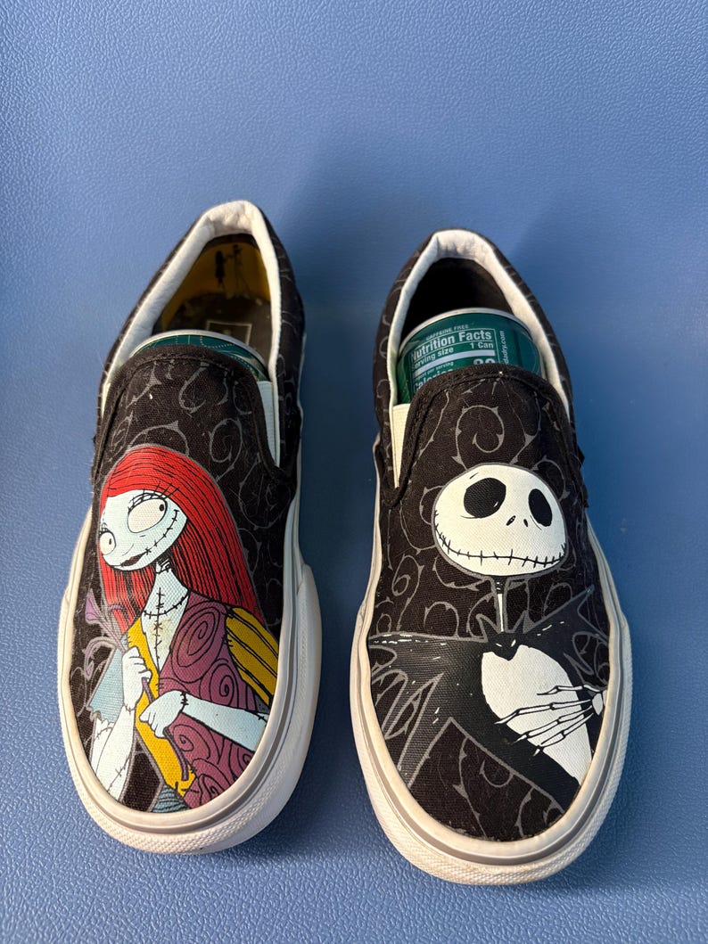 May include: Pair of black slip-on sneakers featuring characters from The Nightmare Before Christmas. One shoe displays Sally, the other Jack Skellington. Both shoes have white soles and a black patterned background.