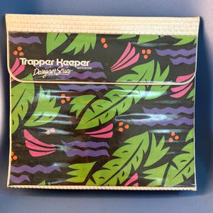 May include: A Trapper Keeper Designer Series notebook with a black background featuring a repeating pattern of green leaves, pink curved shapes, purple wavy lines, and orange dots. The notebook has a white textured border and flap.