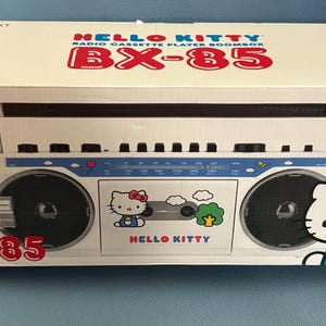 May include: A white rectangular box for a Hello Kitty BX-85 radio cassette player boombox. The packaging features the product name, Bluetooth logo, and graphics of Hello Kitty, clouds, and a tree. The boombox has two black speakers and a blue radio dial.