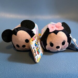 May include: Two plush Disney Tsum Tsum characters, Mickey Mouse and Minnie Mouse, are sitting on a blue surface. Mickey Mouse is wearing a white and pink striped shirt and Minnie Mouse is wearing a white and pink polka dot dress with a pink bow. Both characters have black eyes and black noses.