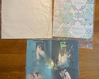 Könnte beinhalten: Drei Packungen Geschenkpapier. Eine ist weiß mit geprägten Mustern, eine zeigt ein blau-grünes Blumenmuster mit dem Text Wedding Wishes und eine zeigt vier blau getönte Fotoszenen eines Paares bei einer Hochzeit.