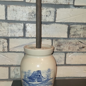 May include: Cream-colored ceramic butter churn with a blue barn scene. The churn has a wooden handle and a lid. The design is reminiscent of vintage farmhouse decor. The churn is approximately 7 inches tall.