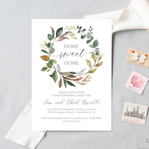 May include: A white invitation card with the text "Home Sweet Home" inside a watercolor wreath of green and brown leaves. The card includes details for a housewarming party, with a roll of white ribbon and decorative stamps.