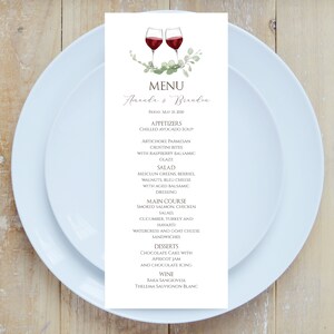Puede incluir: Una tarjeta de menú de boda con una ilustración de acuarela de dos copas de vino y vegetación. El menú incluye aperitivos, ensalada, plato principal, postres y vino. El texto dice "Amanda & Brandon" y "Viernes, 25 de mayo de 2020".