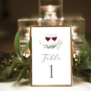 Puede incluir: Una tarjeta de número de mesa con marco dorado y fondo blanco. La tarjeta presenta una ilustración de acuarela de dos copas de vino con vino tinto y hojas verdes. El texto "Mesa 1" está escrito en cursiva.