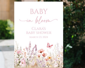 May include: A baby shower sign on a wooden easel. The sign displays the text "BABY in bloom" and "Clara's BABY SHOWER MARCH 23, 2024" in pink script. A watercolour floral design with pink and purple flowers and a butterfly decorates the bottom.