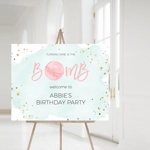 May include: A welcome sign for a birthday party. The sign is on a wooden easel. The sign has a light blue watercolor background with gold flecks. The text reads "Turning nine is the bomb" and "Welcome to Abbie's Birthday Party."