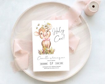 May include: A white invitation card with a watercolor illustration of a cow wearing a flower crown and a pink bow. The text "Holy Cow!" is above the text "Camille is turning one" and event details.