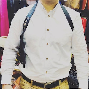 May include: A white button-down shirt with gold buttons and a black leather shoulder strap with silver studs. The shirt is tucked into khaki pants with a black belt.