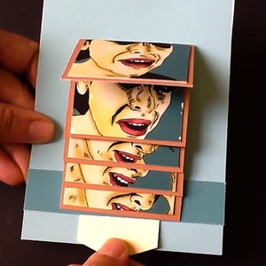 May include: A pop-up card with a layered design featuring a woman's face with different expressions. The card is made of paper and has a blue background.
