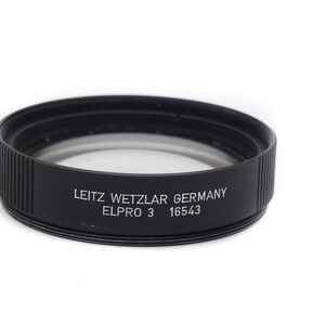 Op de afbeelding: Zwarte metalen filterring met de tekst "LEITZ WETZLAR GERMANY ELPRO 3 16543" gegraveerd aan de zijkant.