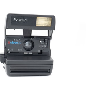 Peut inclure: Un appareil photo instantané Polaroid noir avec un flash intégré et un accessoire d'objectif gros plan. L'appareil photo a un objectif argenté et une étiquette "gros plan" noire et bleue.