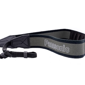 Può includere: Un cinturino per fotocamera nero e grigio con la scritta "Panasonic" stampata in bianco sul cinturino.