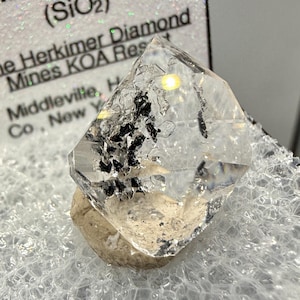 Puede incluir: Un cristal de diamante Herkimer transparente con inclusiones negras, apoyado sobre una pequeña base beige. El cristal está facetado y refleja la luz, mostrando su claridad. El texto de fondo dice "Herkimer Diamond Mines KOA Res".
