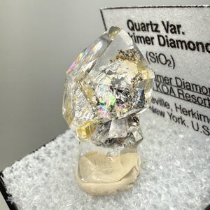 May include: A clear quartz crystal, labelled as Herkimer Diamond, with internal inclusions and iridescent reflections. The crystal is mounted on a white base, with a label that reads "Quartz Var. Herkimer Diamond (SiO2) KOA Resort, New York, U.S.A."