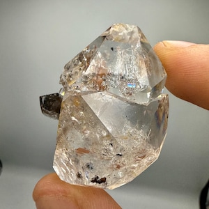 Könnte beinhalten: Ein klarer, facettierter Quarzkristall mit inneren Rissen, kleinen dunklen Mineraleinschlüssen und einer subtilen Regenbogenbrechung. Der Stein weist eine geometrische, kantige Struktur mit scharfen Kanten und ein glasartiges Aussehen auf.