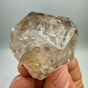 May include: A clear, faceted quartz crystal with a light brown hue. The crystal is held in a hand, showcasing its geometric shape and natural clarity. The surface has a rough texture with internal fractures.