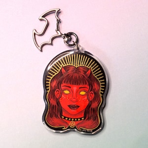 May include: A red and yellow acrylic keychain featuring a cartoon illustration of a woman with horns, a pentagram necklace, and a halo. The keychain is attached to a silver bat-shaped charm.
