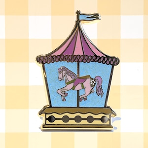May include: A carousel pin featuring a pink horse inside a blue enclosure, topped with a purple and pink tent. The pin has a gold border and a small flag at the top. The horse has a purple saddle and a white flower.