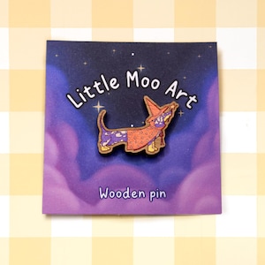 May include: A wooden pin featuring a cartoon dachshund wearing a wizard hat and robe. The pin is against a purple and blue background with stars and the text "Little Moo Art Wooden pin".