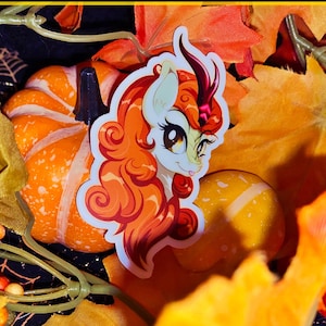 May include: A sticker featuring a cartoon unicorn with orange hair and a horn, set against a backdrop of pumpkins and fall foliage. The sticker has a white background and is surrounded by orange and yellow leaves, creating a seasonal theme.