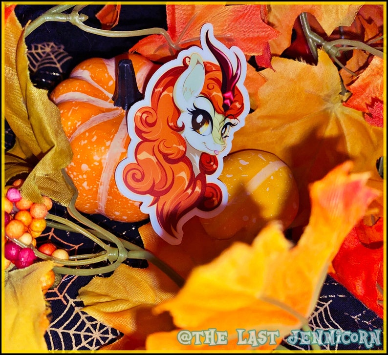 May include: A sticker featuring a cartoon unicorn with orange hair and a horn, set against a backdrop of pumpkins and fall foliage. The sticker has a white background and is surrounded by orange and yellow leaves, creating a seasonal theme.