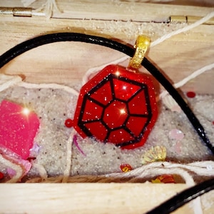 May include: A red pendant with black geometric lines, displayed in a wooden box with sand and decorative elements. The pendant hangs from a black cord and a gold-colored ring.