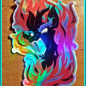 May include: Holographic sticker featuring a black creature with a fiery mane and a fierce expression. The sticker has a rainbow sheen and is surrounded by flames in shades of red, orange, and blue. Text at the bottom reads "@THE LAST JENNICORN".