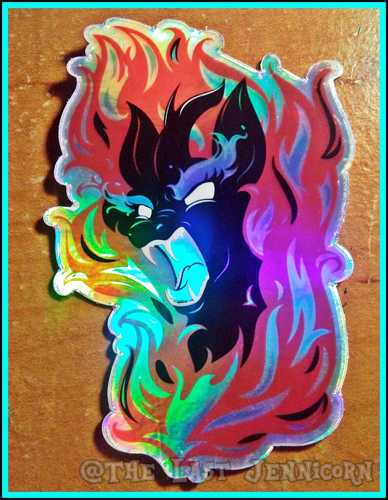 May include: Holographic sticker featuring a black creature with a fiery mane and a fierce expression. The sticker has a rainbow sheen and is surrounded by flames in shades of red, orange, and blue. Text at the bottom reads "@THE LAST JENNICORN".