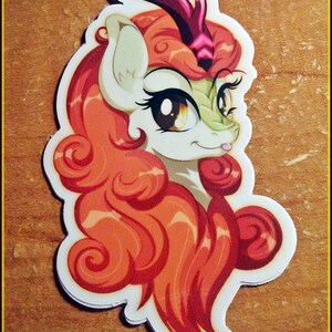 May include: A sticker featuring a cartoon pony with orange and red curly hair, a green face, and large brown eyes. The pony has a white body and a purple horn. The sticker has a white border and the text "@THE LAST JENNICORN" at the bottom.