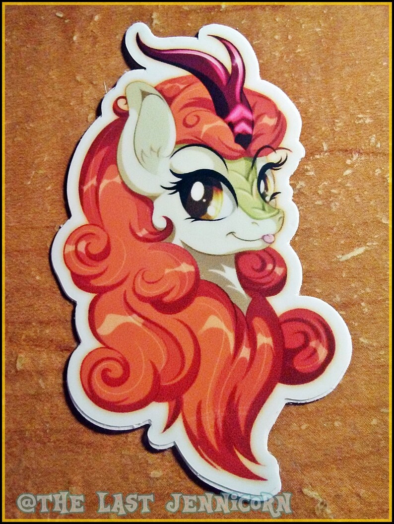 May include: A sticker featuring a cartoon pony with orange and red curly hair, a green face, and large brown eyes. The pony has a white body and a purple horn. The sticker has a white border and the text "@THE LAST JENNICORN" at the bottom.