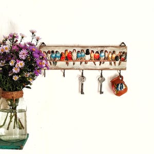 May include: A wooden key holder with a colorful bird design. The holder has five hooks and is decorated with a row of birds on a wire. There are two keys and a small brown jug hanging from the hooks.
