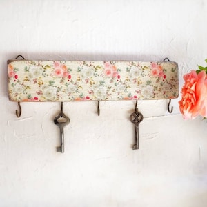 May include: A wooden key holder with a floral pattern in shades of pink, white, and green. It has five hooks for hanging keys.