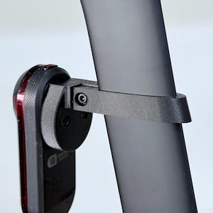Könnte beinhalten: Ein schwarzes Fahrradrücklicht ist mit einem schwarzen Riemen und einer Halterung an einer schwarzen Sattelstütze befestigt. Das Rücklicht hat eine rote Linse und ein schwarzes Gehäuse. Die Sattelstütze ist dunkelgrau.