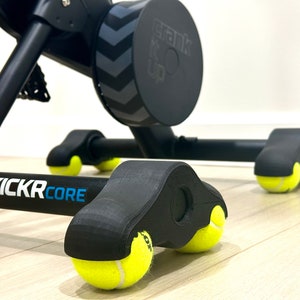 以下が含まれることがあります： 黒い屋内サイクルトレーナーのクローズアップ。フレームに「KICKRCORE」の文字が印刷されています。トレーナーには、黄色いテニスボールが取り付けられた車輪が4つあります。