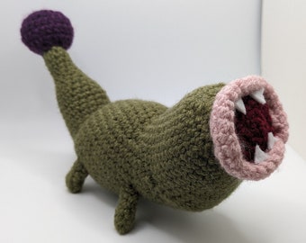 May include: A crocheted plush toy shaped like a green monster with a large open mouth, pink teeth, and a purple ball on top of its head.