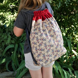 May include: A child wearing a grey shirt and white shorts is carrying a red and beige drawstring backpack with a monkey pattern.