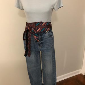 May include: A light blue short-sleeved ribbed t-shirt paired with blue denim jeans featuring a wide cuff at the hem. A long, wrap-around fabric belt with a dark maroon, teal, and orange patterned design is tied around the waist of the jeans.