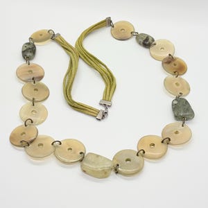 May include: A necklace featuring multiple circular beige horn-like discs and irregular green stone beads connected by metal jump rings. The necklace includes a multi-strand olive green suede cord section with a silver-toned metal clasp closure.
